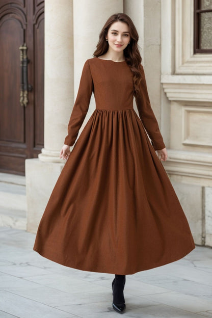 A woman wearing a brown maxi winter dress with pleated detail at the waist, long sleeves, and side seam pockets.