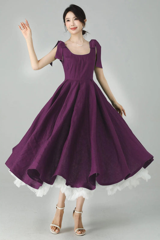 May include: A plum-colored dress with a square neckline and short sleeves featuring bow details. The dress has a full skirt with a white lace trim at the hem. The model is wearing white sandals.