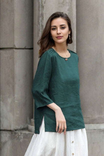 May include: A green linen top with a round neckline and three-quarter sleeves. The top has a side slit on the left side. The top is paired with a white skirt.