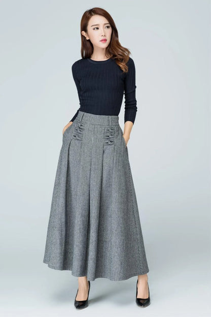May include: A black long-sleeved ribbed sweater paired with a high-waisted grey houndstooth midi skirt. The skirt features a wide waistband, side pockets, and decorative stitched pleat details at the front, falling in a full A-line silhouette.