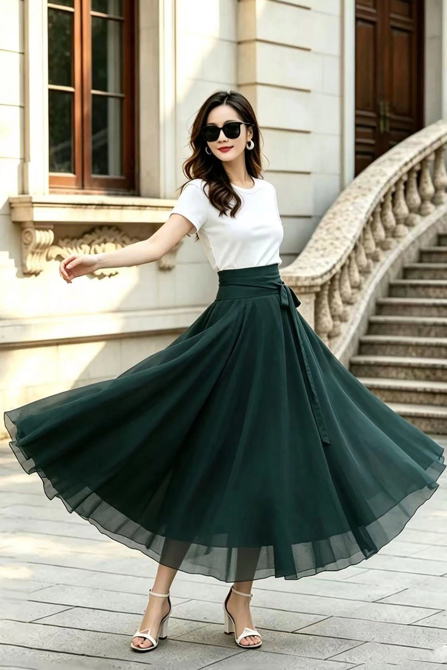 May include: A white short-sleeved t-shirt paired with a high-waisted, floor-length dark green skirt. The skirt features a wide waistband with a tie detail and a full, flowing silhouette made of a sheer, layered fabric that flares outward.