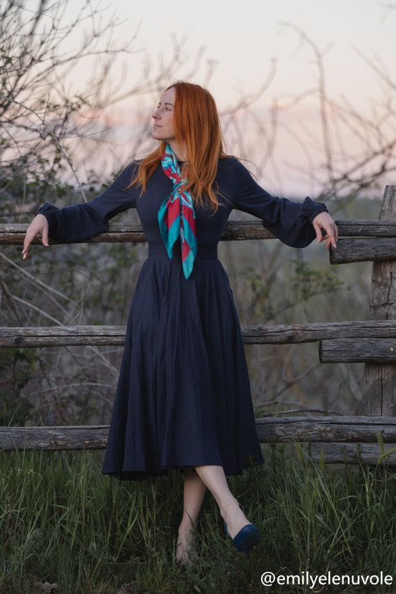 Woman in a long dark dress with a colorful scarf standing by a wooden fence in a natural setting.