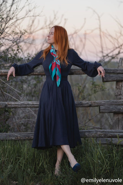 Woman in a long dark dress with a colorful scarf standing by a wooden fence in a natural setting.