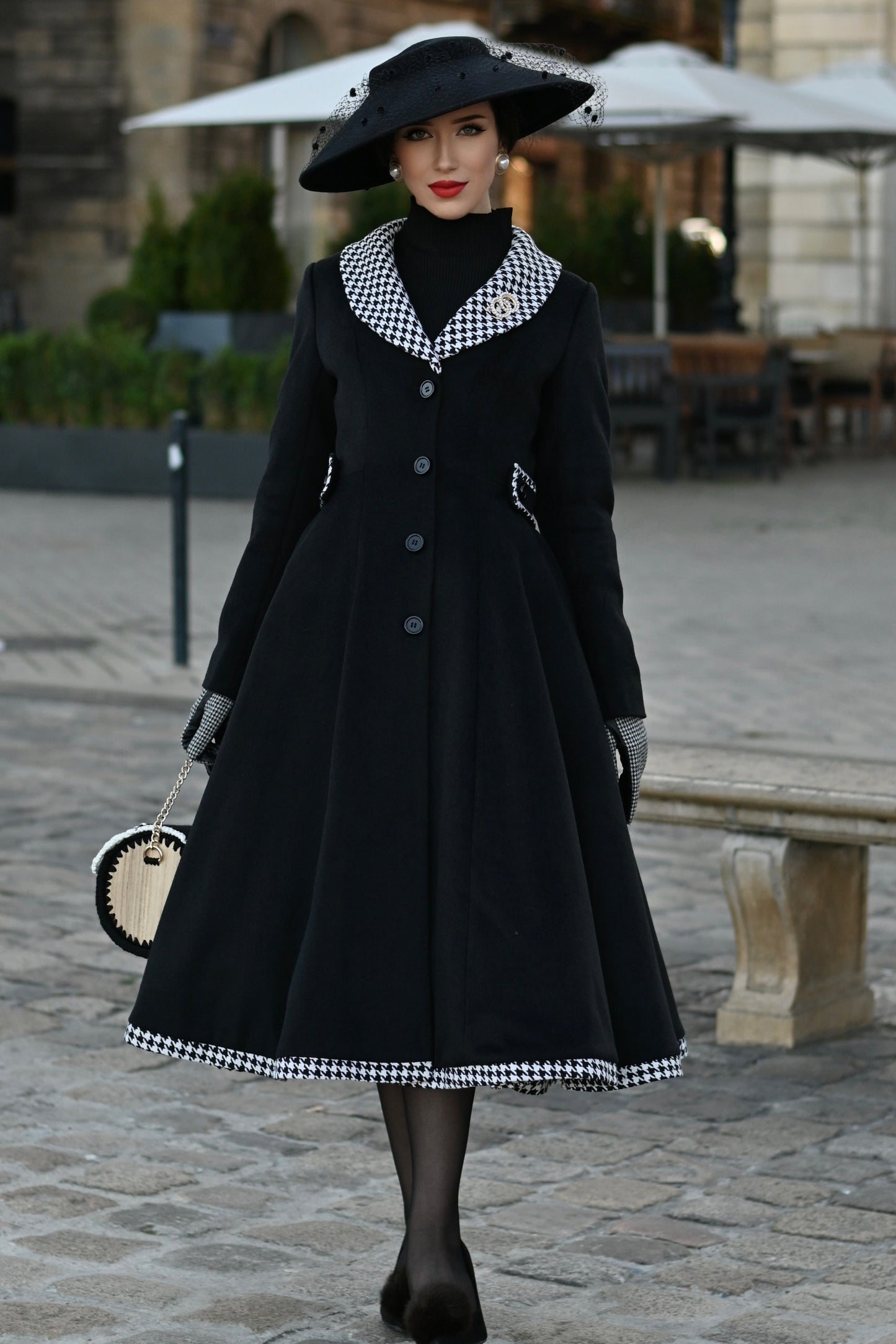 Woman in a black coat and hat standing on a street with buildings and tables in the background.