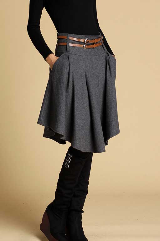 A model wearing a dark gray pleated wool mini skirt with a wide waistband, side pockets, and an asymmetrical hemline. The skirt is paired with a black turtleneck and knee-length boots.