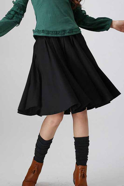 A woman wearing a green top with three-quarter sleeves and a black pleated midi skirt with a wild waistband. She is also wearing brown ankle boots.