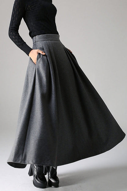 A woman modeling a long maxi wool skirt in gray, with a fitted waist and pleated detail at the waist, standing against a plain background.