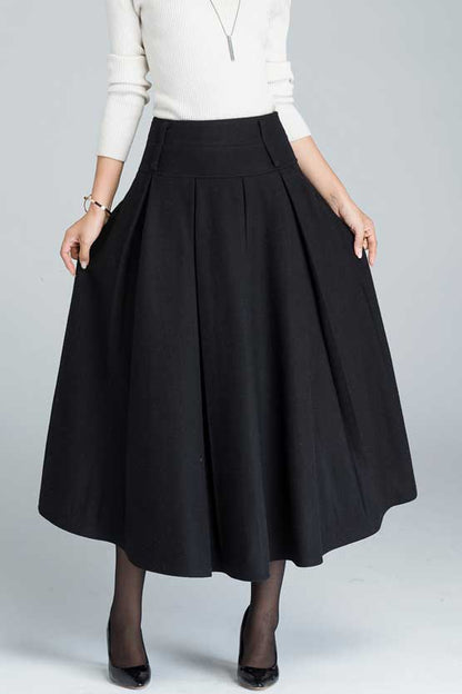 A woman wearing a dark gray pleated midi skirt with a wide waistband and twin side pockets, paired with a black top and black shoes. Accessory belt with a brown buckle is visible.