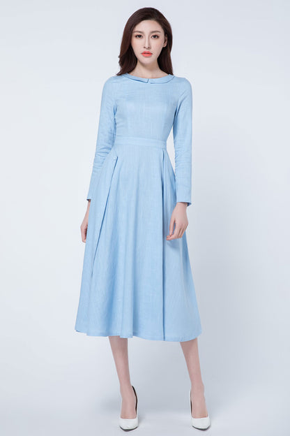 A woman standing wearing a light blue pleated midi dress with a round-neckline and small lapel collar, featuring a fitted waist and long sleeves.