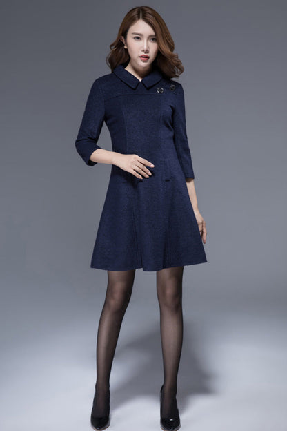 A navy blue wool tunic dress with a lapel collar, fitted waist, and A-line skirt, featuring button decorations and a back zipper closure. The model is posing against a grey background.
