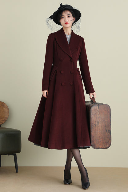 A woman standing in a wine red, medium-weight, double-breasted wool coat with button closure and a wide-skirted design, holding a suitcase and wearing a black hat and black shoes.