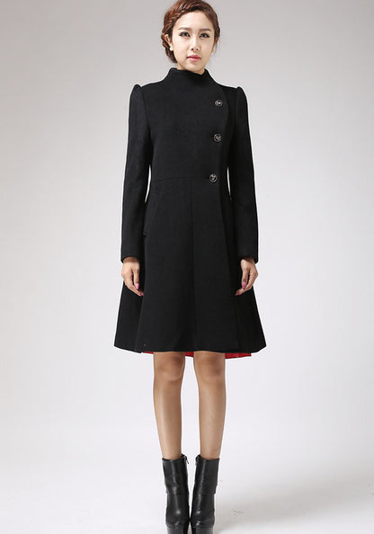 A woman standing wearing a black winter coat with asymmetric button closure and a high stand-up neckline. The coat has long sleeves and is paired with black boots.