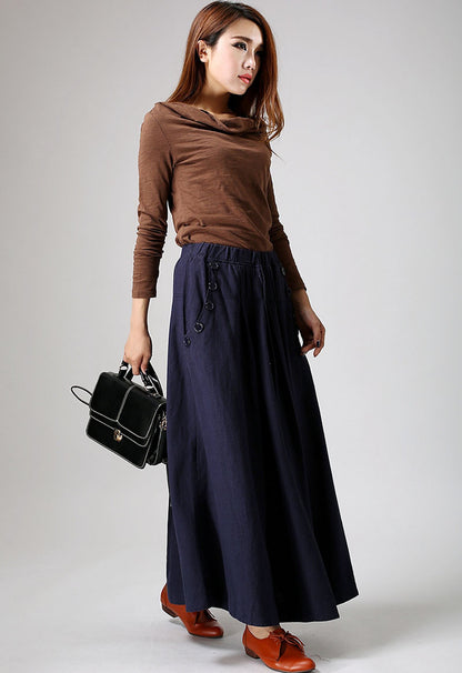 A woman wearing a brown top and blue linen maxi skirt with pockets, carrying a black bag and wearing red shoes.