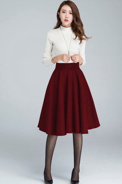 A high-waisted, A-line wool circle skirt in red, with a visible blue stripe wool blend and a pleated detail at the waist. The model is wearing a white sweater and black shoes.
