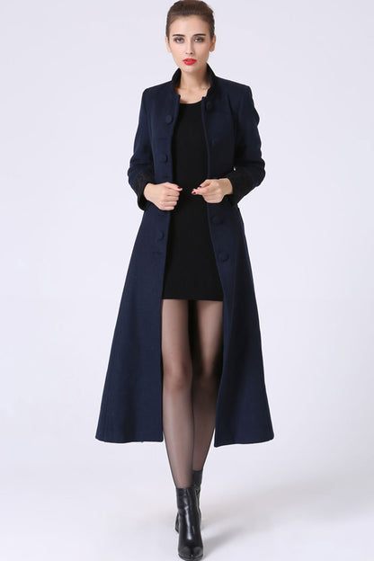 A woman standing wearing a long blue wool coat with a mandarin collar neckline, hip side pockets, and a tie belt waist, paired with a black dress and black boots.