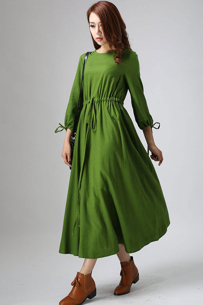 A woman wearing a green maxi dress with 3/4 sleeves and a round neckline, standing in a room with a brown bag and tan shoes.