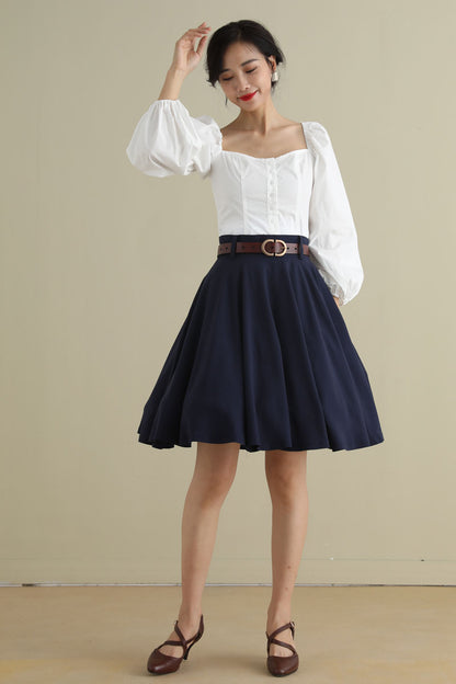 A woman wearing a navy blue high-waisted full circle skirt with pockets and a white top, posing against a plain background.