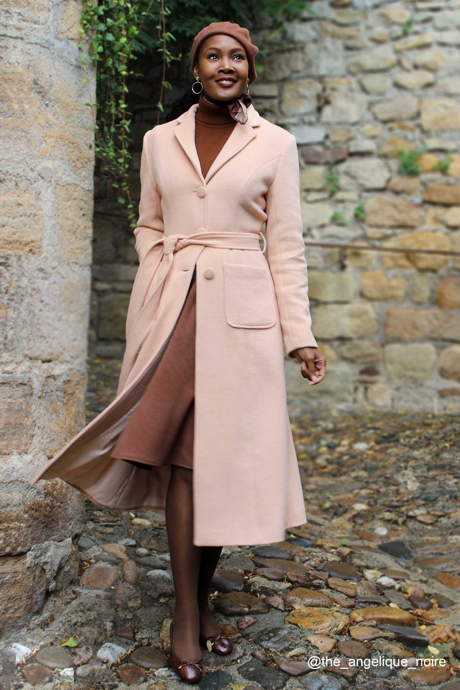 A woman standing outdoors wearing a long camel brown wool coat with a single breasted design and a self-tie belt waist. She also wears a brown hat and dark shoes.