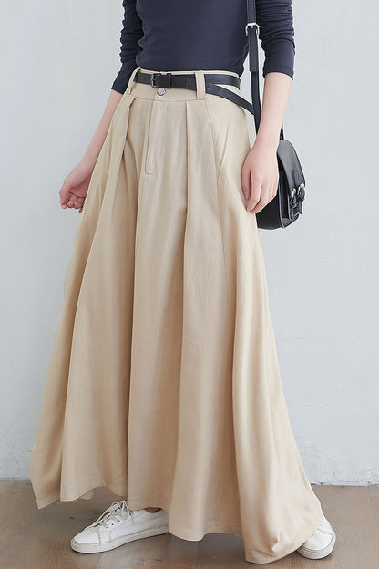 A woman wearing a beige maxi linen skirt with a black top and white sneakers. The skirt features a front zipper and buttons closure, side pockets, and contrast patchwork details.