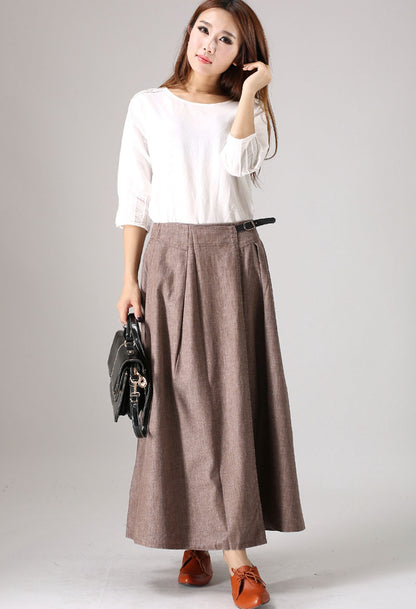 A woman wearing a white top and a brown linen maxi skirt with pleats at the waist, holding a black bag and standing against a neutral background.