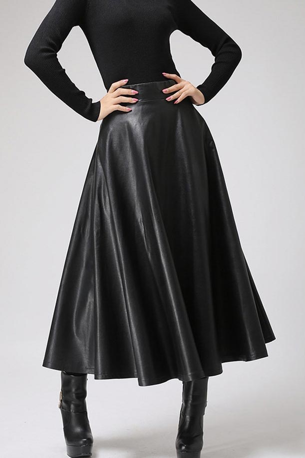 A person wearing a black faux leather maxi skirt with a black long-sleeve top and black boots, standing against a grey background.