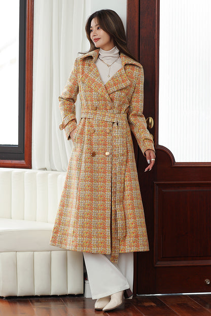 A woman standing in a doorway wearing a yellow and blue plaid wool coat with a V-neckline, long sleeves, and a belt.