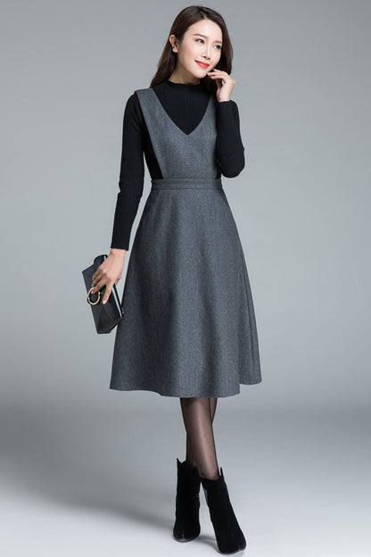 A model wearing a gray wool sleeveless midi skirt with suspenders, featuring two pockets on each side and a side zipper closure. She is also wearing a black top and black ankle boots.