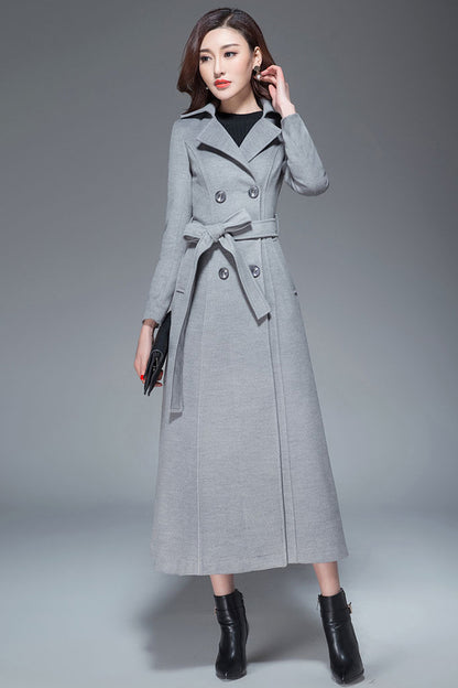 A woman wearing a long, double-breasted wool coat with a belt, button closure, and side pockets. The coat is gray and the model is also wearing black boots and a black bag.