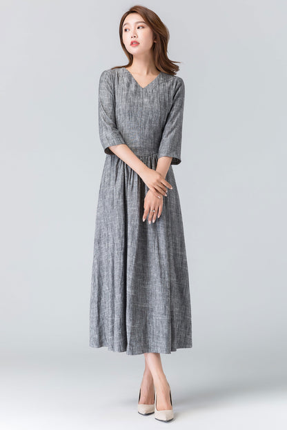 A woman standing, wearing a gray linen pleated midi dress with a V-neckline and 3/4 length sleeves.