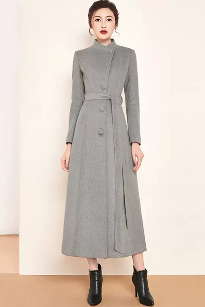 A woman standing wearing a long, medium-weight gray wool coat with a belt, button closure, and two side pockets. She has a fit and flare silhouette and is wearing black boots.