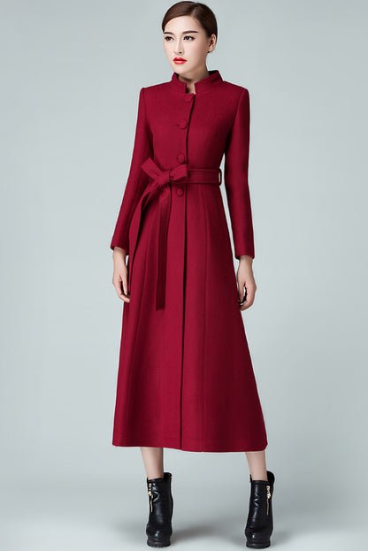 A woman standing wearing a red maxi warm wool coat with a belt, button closure, and two side pockets. The coat has a fit and flare silhouette.