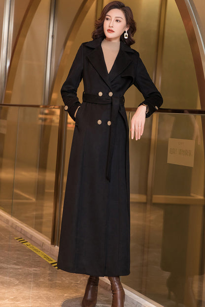 A woman standing in a lobby wearing a long black wool coat with double-breasted button closure, a belt, and two side pockets. She has a subtle, elegant presence.
