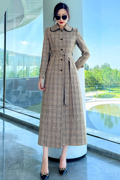 A woman standing in a modern space wearing a long plaid handmade wool coat in a brown and beige color, with a belt and button closure, paired with sunglasses and high-heeled shoes.