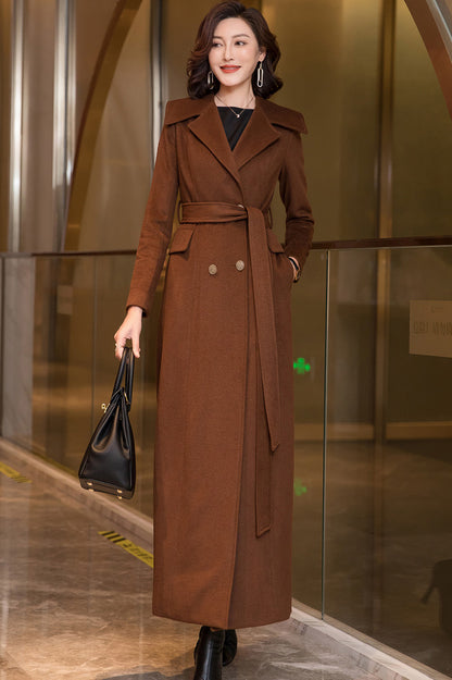 A woman standing in a lobby wearing a long double-breasted wool coat in brown, with a belt and button closure, paired with black boots and a black bag.
