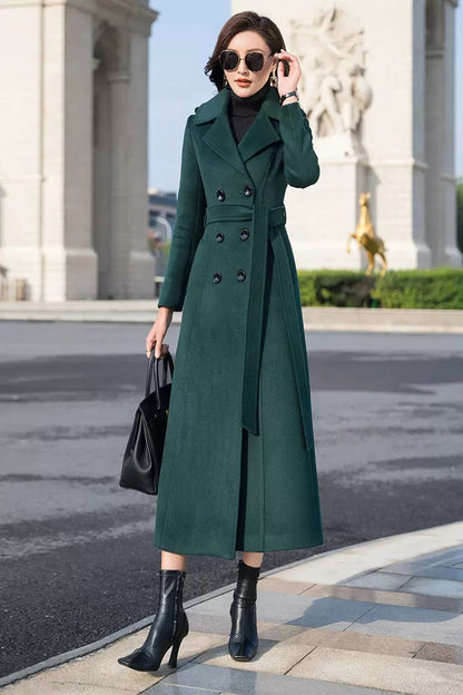 A woman standing on a pavement wearing a long green handmade wool coat with a belt, black boots, sunglasses, and a black bag.