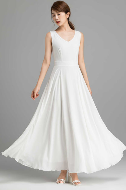 A woman modeling a white chiffon maxi dress with a V neckline and back zip closure, standing on a plain background.