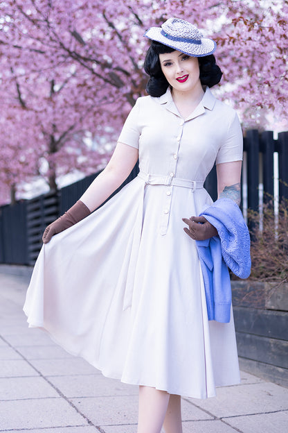 A woman wearing a beige 50s swing A-line handmade shirt dress with short sleeves, a notched collar, and front button-up closure, posing outdoors with pink cherry blossoms in the background.